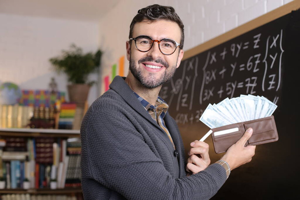 close-up portrait of handsome young teacher holding wallet full of cash in front of blackboard in classroom - Φωτογραφία, εικόνα