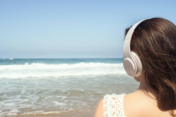 Alone woman on a beach in headphones listen music looking on the sea. Female relaxation at summer vacation. Back view - Photo, image