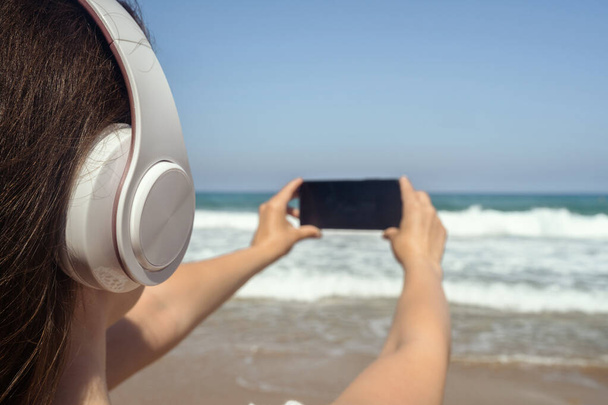 Alone woman on a beach in headphones listen music, taking picture of sea using phone. Female relaxation at summer vacation. Back view - Photo, Image