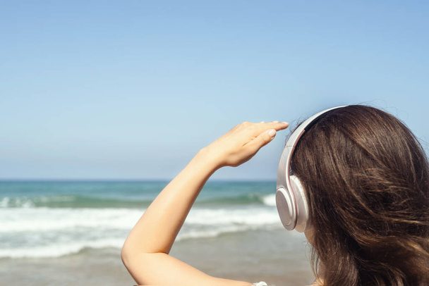 Allein am Strand hört eine Frau mit Kopfhörern Musik, die auf das Meer blickt. Weibliche Entspannung in den Sommerferien. Rückseite - Foto, Bild