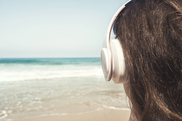 Alone woman on a beach in headphones listen music looking on the sea. Female relaxation at summer vacation. Back view - Zdjęcie, obraz