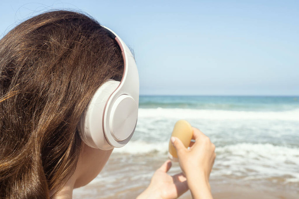 Woman in headphones listen music applying sunscreen on skin  on a beach looking on the sea. Female relaxation at summer vacation. Back view - Fotó, kép