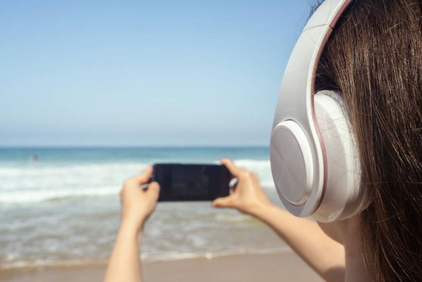 Alone woman on a beach in headphones listen music, taking picture of sea using phone. Female relaxation at summer vacation. Back view - 写真・画像