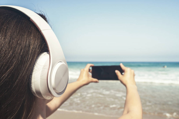 Alone woman on a beach in headphones listen music, taking picture of sea using phone. Female relaxation at summer vacation. Back view - 写真・画像