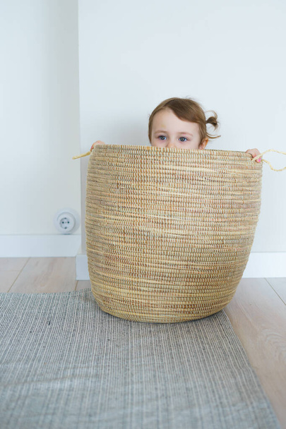 little girl with two ponytails blonde hair in light leggings and a beige T-shirt plays at home with objects,toys, hides in a wicker basket plays on the bed, bright white apartment,draws flowers - Foto, Imagem