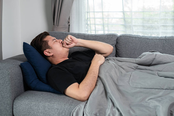 Asian man is lying with blanket on couch at home to rest when having high fever, headache and using hand cover your mouth when cough and sneezing - Zdjęcie, obraz