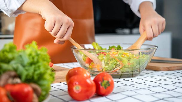 Close up of asian housewife wearing apron and using ladle to mixing vegetable salad in bowl while standing preparing to cooking healthy meal for breakfast, lunch and dinner in the kitchen at home - Fotó, kép