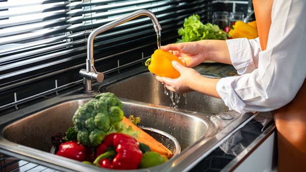 Κοντινό πλάνο της ασιάτισσας νοικοκυράς φορώντας ποδιά για το πλύσιμο πιπεριού με νερό και λαχανικά σε σουρωτήρι στο νεροχύτη για την προετοιμασία για να κάνει υγιεινό γεύμα στην κουζίνα στο σπίτι - Φωτογραφία, εικόνα