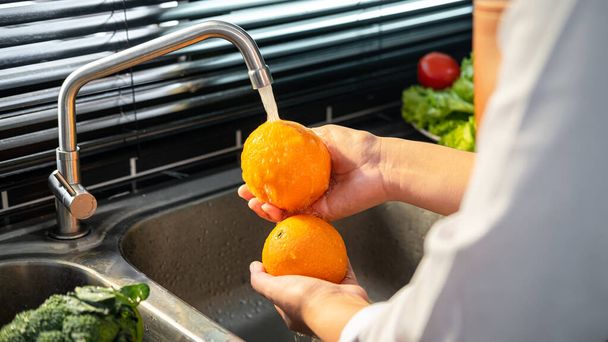 Hands of asian housewife washing orange and vegetables with water in sink while wearing apron and preparing to cooking for healthy meal in the kitchen at home - Фото, изображение