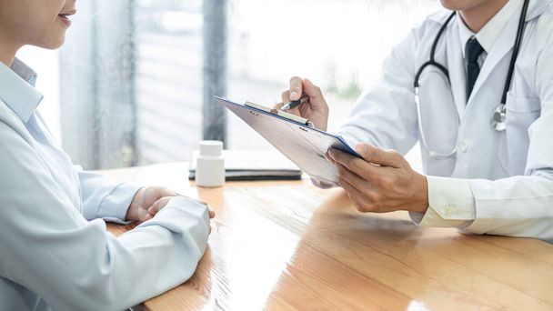 Male doctor with stethoscope is writing symptom of patient medication history and prescription on clipboard while explaining treatment and giving recommendation to patient in clinic - Foto, Imagem