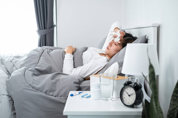 Asijská žena pocit nevolnosti a spí v přikrývce na posteli, zatímco drží tkáň v ruce a dotýká se čela po použití teploměru pro kontrolu tělesné teploty a vzít léky v ložnici doma - Fotografie, Obrázek