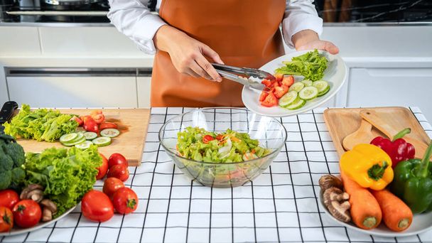 Azjatycka gospodyni domowa trzyma talerz z sałatą, ogórkiem i pomidorami i używa szczypiec warzywnych do miski do mieszania sałatki na lunch podczas noszenia fartucha i stojąc do gotowania zdrowego posiłku w kuchni - Zdjęcie, obraz