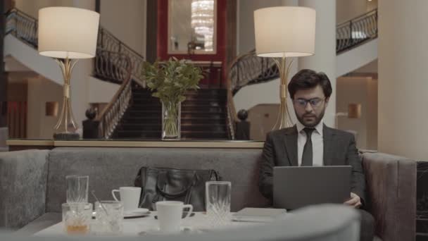 Medium long of young man wearing grey formal suit and glasses, using laptop computer, then talking to unrecognizable person joining him across table in business center lobby - Footage, Video
