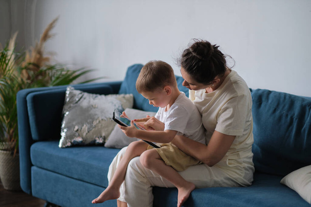madre e hijo con la tableta en casa. Madre mostrando contenido multimedia en línea a su hijo en una tableta sentada en un sofá en la sala de estar en el interior de una casa - Foto, imagen