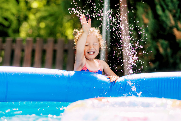Ένα χαριτωμένο κοριτσάκι ρίχνει νερό σε μια φουσκωτή πισίνα στον κήπο και γελάει. Καλοκαίρι, νερό, σπρέι, παιδιά - Φωτογραφία, εικόνα