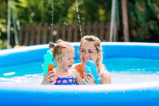 Mladá matka a její roztomilé dítě si hrají v bazénu s vodní pistole, stříkající vodu kolem - Fotografie, Obrázek