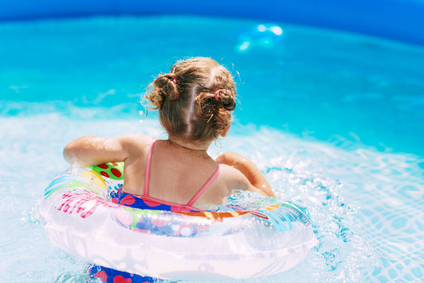 Uma menina encantadora em um maiô brilhante aprende a nadar na piscina no jardim com a ajuda de um círculo inflável. Hora de verão, recreação, visão traseira - Foto, Imagem