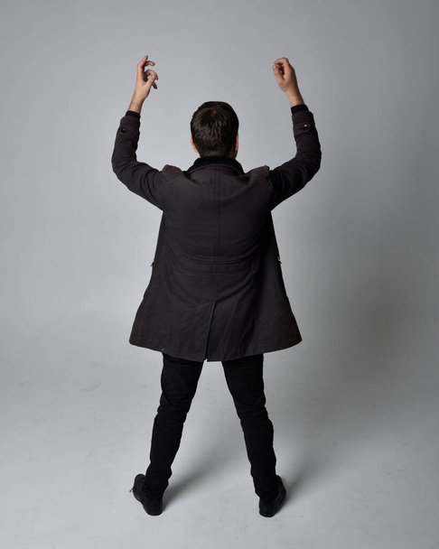 Full length portrait of a brunette man wearing black leather coat.  Standing pose facing backwards, walking away from the camera isolated  against a grey studio background. - Foto, afbeelding