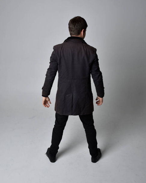 Ολόκληρο πορτραίτο ενός μελαχρινού άντρα που φορούσε μαύρο δερμάτινο παλτό. Στέκεται στάση κοιτάζοντας προς τα πίσω, περπατώντας μακριά από την κάμερα απομονωμένη σε ένα γκρι φόντο στούντιο. - Φωτογραφία, εικόνα
