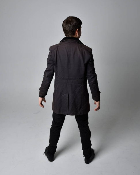 Volledig portret van een brunette met een zwarte leren jas. Staande houding naar achteren gericht, weglopen van de camera geïsoleerd tegen een grijze studio achtergrond. - Foto, afbeelding
