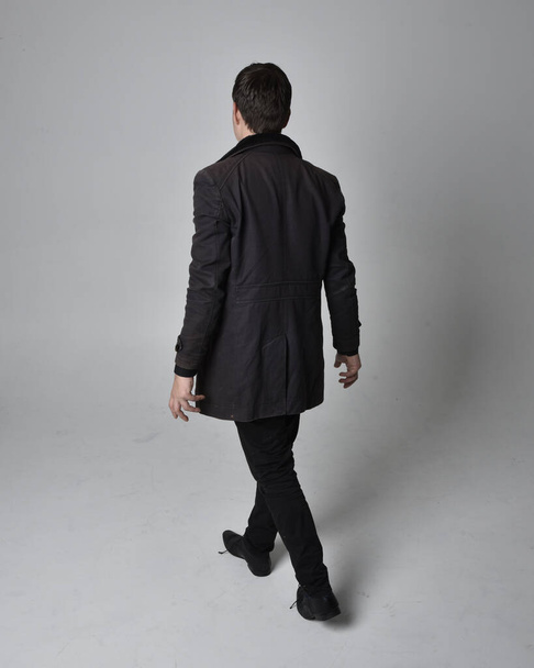 Retrato de comprimento total de um homem morena usando casaco de couro preto. Posição de pé virada para trás, afastando-se da câmera isolada contra um fundo de estúdio cinza. - Foto, Imagem