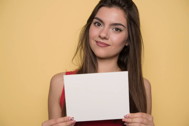 美しい若い女性が黄色い背景に白紙を持っていて - 写真・画像