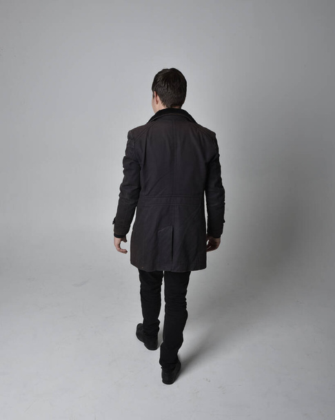 Full length portrait of a brunette man wearing black leather coat.  Standing pose facing backwards, walking away from the camera isolated  against a grey studio background. - Fotó, kép