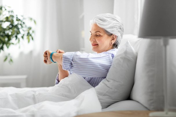 happy old woman with health tracker sitting in bed - Zdjęcie, obraz