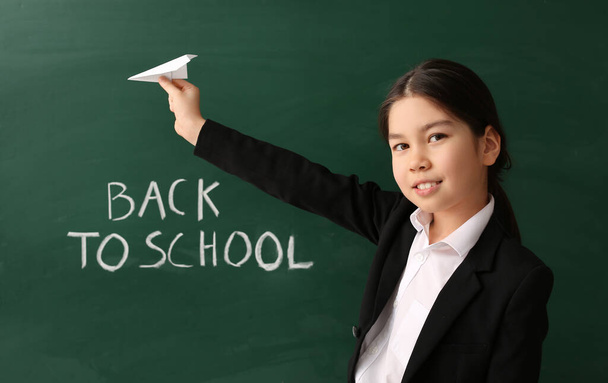 Schattig Aziatisch schoolmeisje met papier vliegtuig in de buurt van schoolbord in de klas - Foto, afbeelding