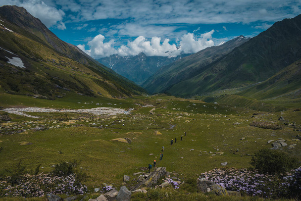 Group of people trekking in a beautiful vast mountain valley with white rhododendrons under a partially open sky with beautiful cloud patterns while doing the Bali pass trek in the Sankri region of Garhwal Himalayas in Uttarakhand.  - Foto, imagen