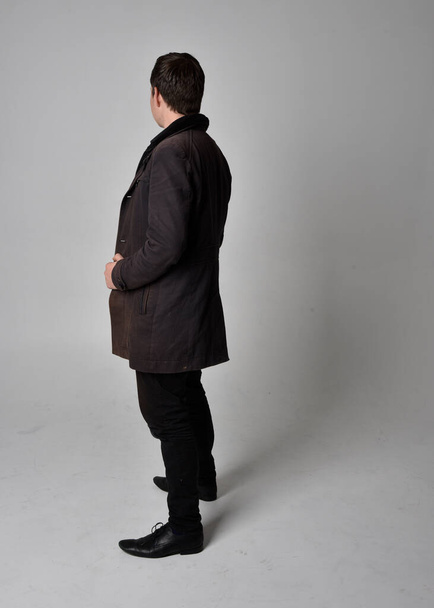 Портрет брюнетки в чорному шкіряному пальто. Позування стоячи ізольовано на сірому фоні студії
. - Фото, зображення
