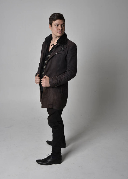 Full length portrait of a brunette man wearing black leather coat.  Standing pose  isolated  against a grey studio background. - Photo, image