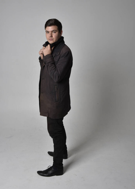 Ganzkörperporträt eines brünetten Mannes in schwarzem Ledermantel. Standing Pose isoliert vor grauem Studiohintergrund. - Foto, Bild