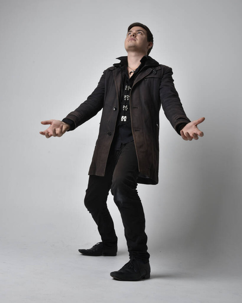 Full length portrait of a brunette man wearing black leather coat.  Standing pose  isolated  against a grey studio background. - Photo, Image