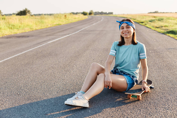 Photo of beautiful smiling female wearing t shirt, hair band and short, sitting on street with longboard, looking at camera with toothy smile, expressing happiness, enjoying leisure time. - Φωτογραφία, εικόνα