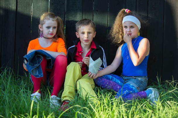 Belas crianças modelo em roupas brilhantes sentar na grama perto de uma cerca de madeira. - Foto, Imagem