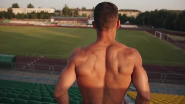 Een jonge man met een naakte romp staat in het stadion en buigt zijn spieren op zijn rug. Langzame beweging - Video