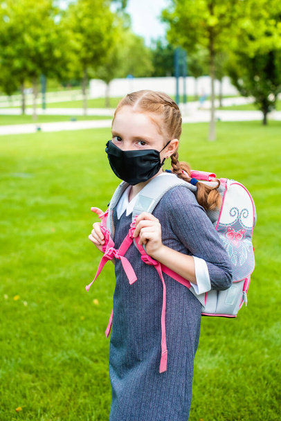 a pretty schoolgirl with red hair and a medical mask goes to school. behind her shoulders is a school bag, on the head there are two pigtails. - Foto, Imagem