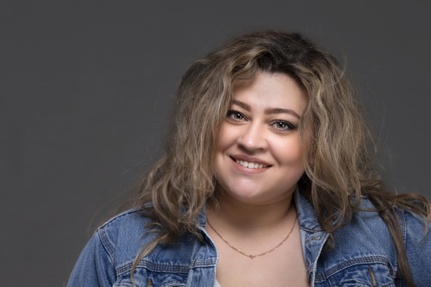 Portrait of a beautiful fat woman in a denim jacket looking at the camera and smiling on a gray background. - Fotografie, Obrázek