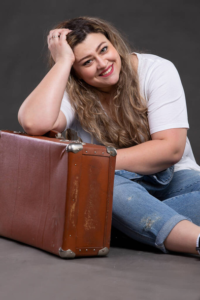Beautiful fat woman in denim clothes with an old retro suitcase on a gray background. Plus size model girl. - Fotó, kép