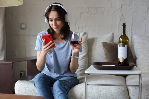 Pretty girl using her smartphone on couch at home in the living room. Listening music, drinking red wine, relaxation after a hard week at work - Foto, Bild