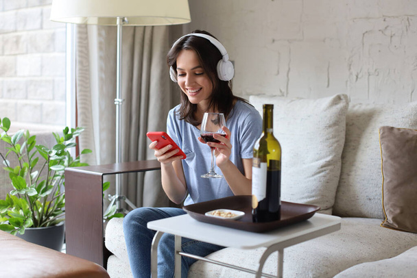 Pretty girl using her smartphone on couch at home in the living room. Listening music, drinking red wine, relaxation after a hard week at work - Foto, Imagen