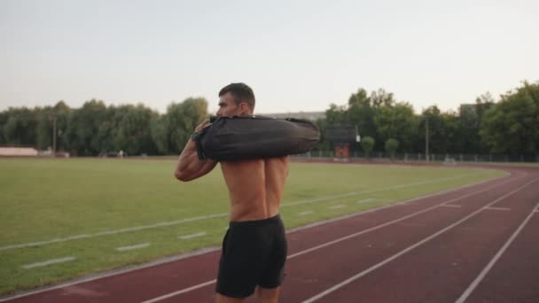 Een bodybuilder in zwarte shorts loopt door het stadion loopband met een zandzak op zijn schouders en kijkt achterom. Langzame beweging - Video