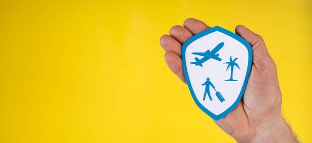 Έννοια της ταξιδιωτικής ασφάλισης με χάρτινη ασπίδα που κρατείται από χέρι σε κίτρινο φόντο - Φωτογραφία, εικόνα