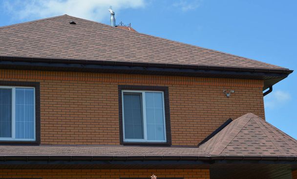 A close-up of a house with an asphalt shingled roofing construction, a roof ventilation cap, plastic soffit, rain gutters, and security cameras installed.  - Photo, Image