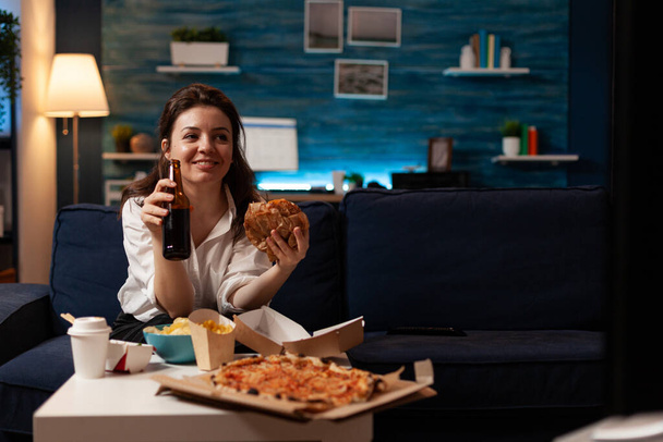 Happy woman holding beer bottle eating tasty takeaway food delivery while watching entertainment movie - Photo, Image
