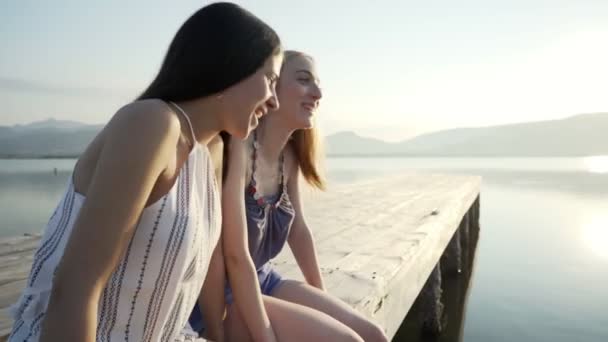Two cute best carefree friends sitting on wooden pier at sunset or dawn with dangling feet in summer clothes talking and joking with each other. 20s girls having fun spending time together in nature - Footage, Video