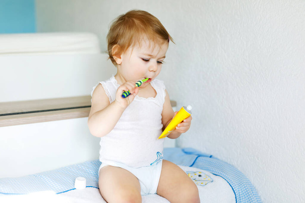 Pikkutyttö pitelee hammasharjaa ja harjaa ensimmäiset hampaat. Pikkulapsi opettelee puhdistamaan maitohampaan. Ennaltaehkäisy, hygienia ja terveydenhuollon käsite. Onnellinen lapsi kylpyhuoneessa - Valokuva, kuva