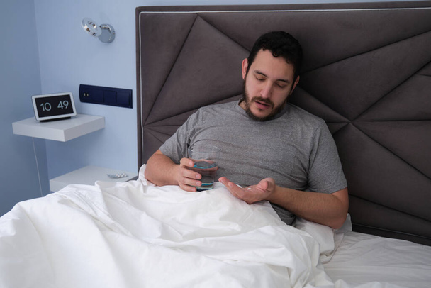 Egy fiatal latin férfi beteg az ágyban, tablettát és egy pohár vizet szed. Rossz közérzet. - Fotó, kép