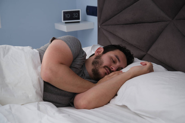 Un joven latino durmiendo en la cama por la mañana. Reloj digital en el fondo. - Foto, imagen